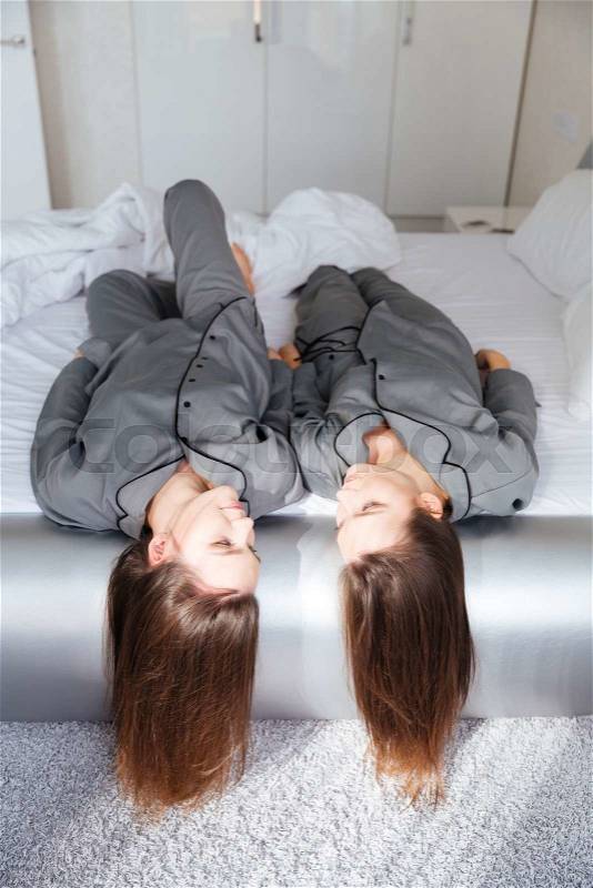 Two charming young sisters twins in grey pajamas lying on bed, stock photo