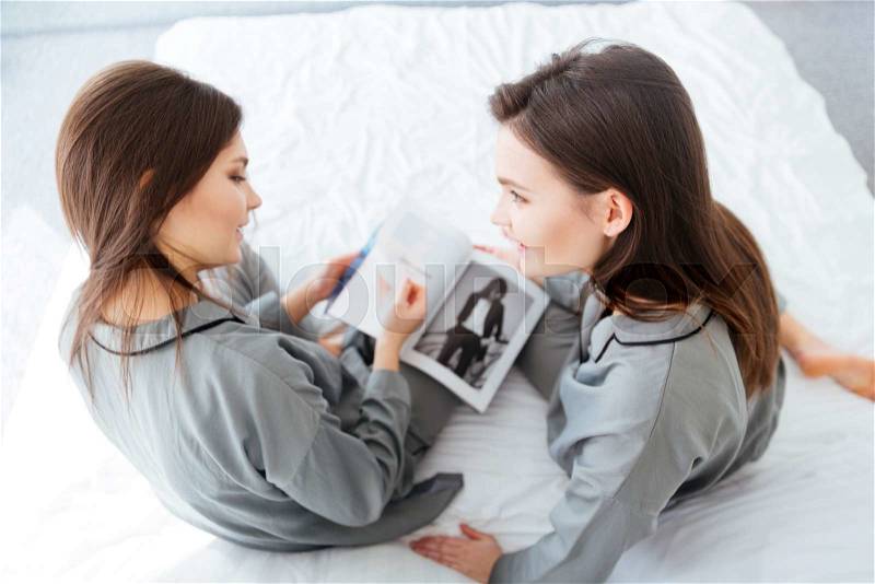 Two attractive young sisters twins talking and reading magazine in bedroom, stock photo