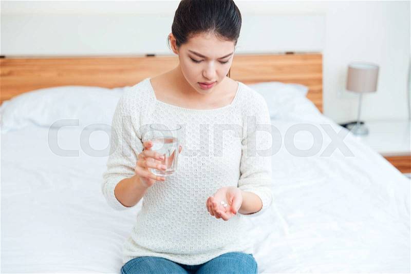 Sick woman holding pills and glass with water on the bed at home, stock photo