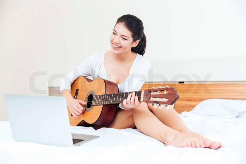 Happy woman playing records on guitar supported by laptop computer on the bed at home, stock photo