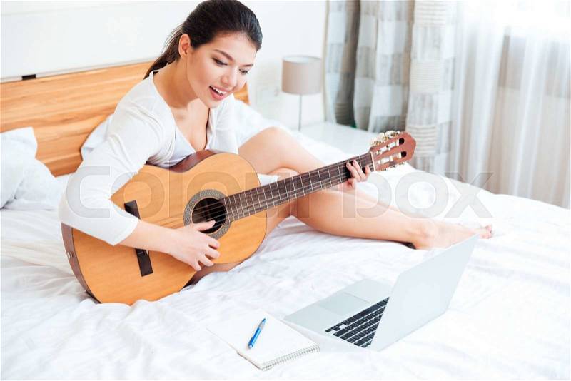 Smiling woman playing records on guitar supported by laptop computer on the bed at home, stock photo