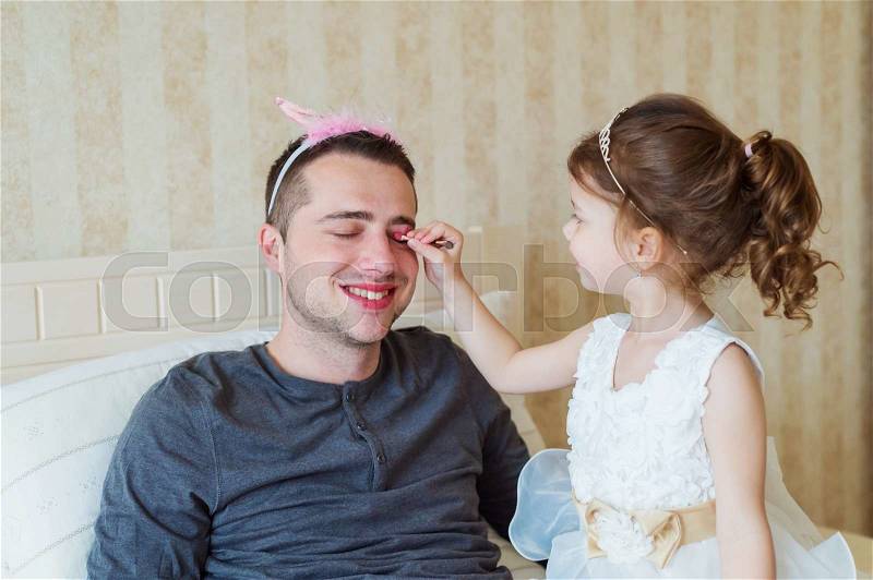 Cute little girl in princess dress putting on colorful make up on her father, stock photo