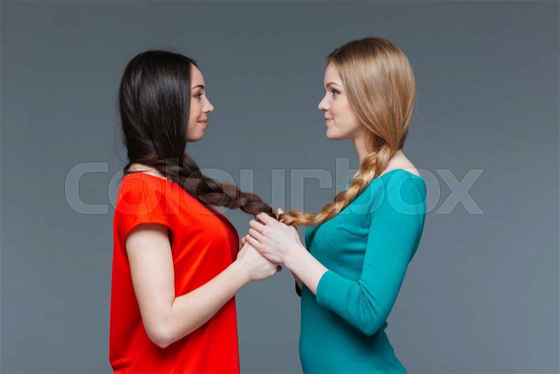 Two smiling cute young women holding hands and looking on each other over grey background, stock photo