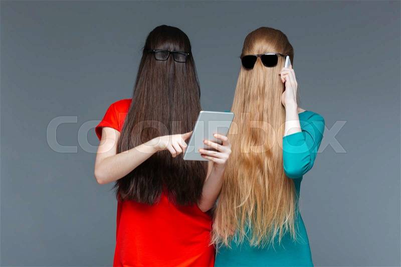 Two amusing young women with faces covered by long hair using tablet and cell phone over grey background, stock photo