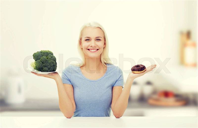Healthy eating, junk food, diet and choice people concept - smiling woman choosing between broccoli and donut over kitchen background, stock photo