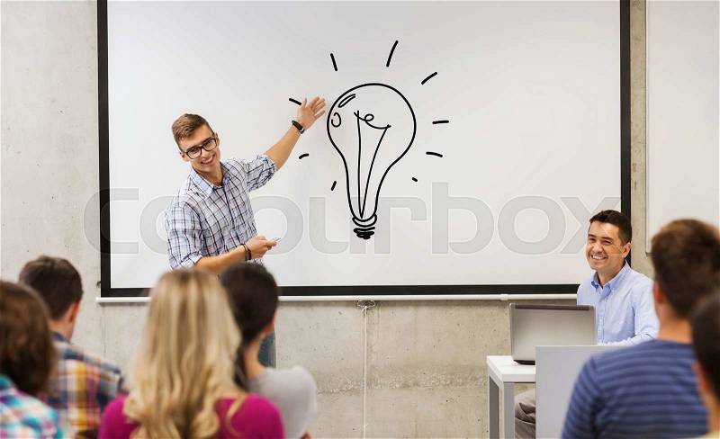 Education, high school, idea and people concept - student standing with remote control in front of teacher and classmates in classroom and showing light bulb on white board in classroom, stock photo
