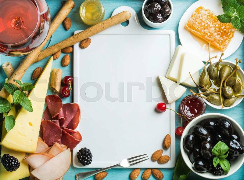 Summer wine snack set. Glass of rose, meat, cheese, olives, honey, bread sticks, nuts, capers and berries with white ceramic board in center, blue wooden background, top view, copy space, stock photo