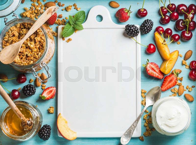 Healthy breakfast ingredients. Oat granola in open glass jar, yogurt, fruit, berries, honey and mint on blue background with white ceramic board in center, top view, copy space, stock photo