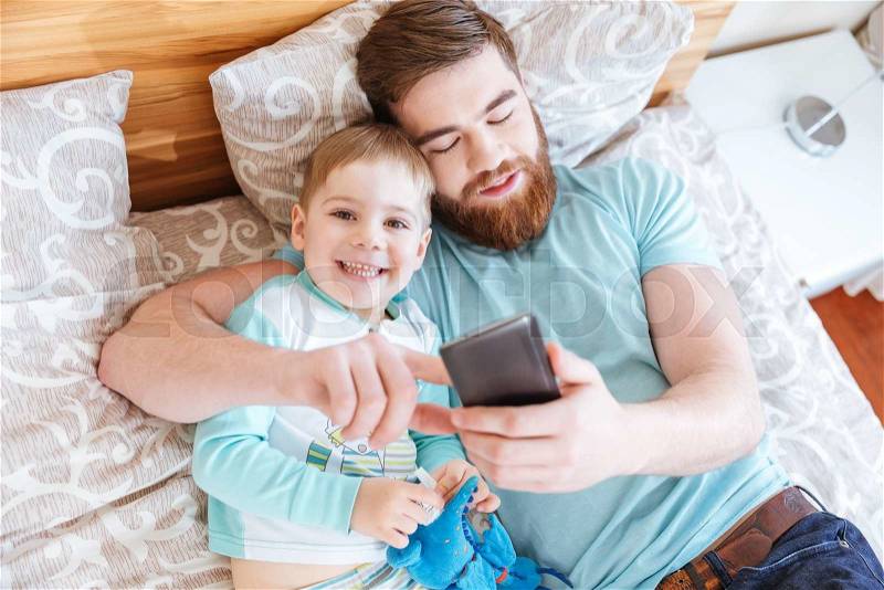 Top view of dad and son lying on bed and using cell phone together, stock photo