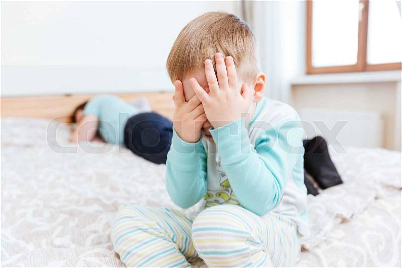 Sad little boy sitting and crying on bed while his father is sleeping, stock photo