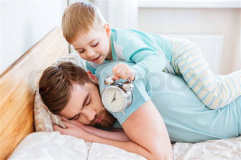 Cute little son holding alarm clock near sleeping father ear at home, stock photo