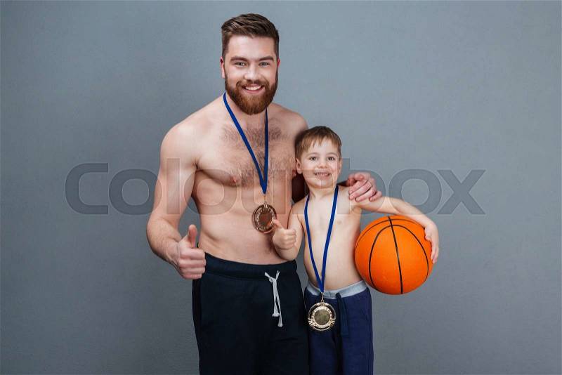 Cheerful man and his son with golden medals and basketball ball showing thumbs up over grey background, stock photo