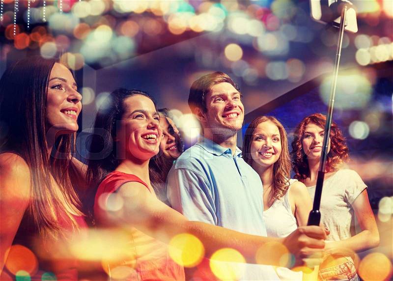 Party, technology, nightlife and people concept - smiling friends with smartphone and monopod taking selfie in night club with holidays lights, stock photo