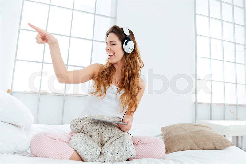 Cheerful woman in pajamas sitting on the bed with tablet computer and pointing finger away, stock photo