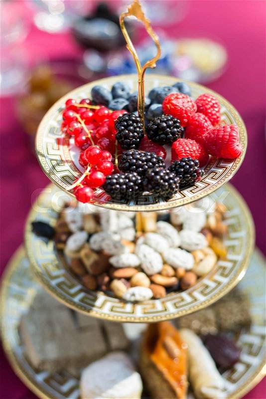 Plate with berries - red current, blackberry, raspberries and and blackberry - in focus and nuts and cakes out of focus on a background, stock photo