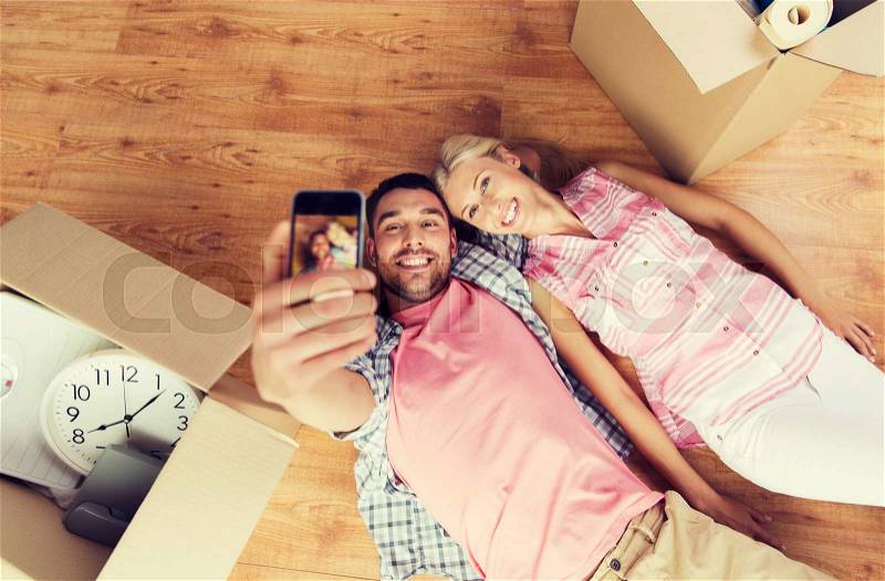 New home, technology, people, repair and moving concept - happy couple taking selfie with smartphone and lying on floor among cardboard boxes at home, stock photo