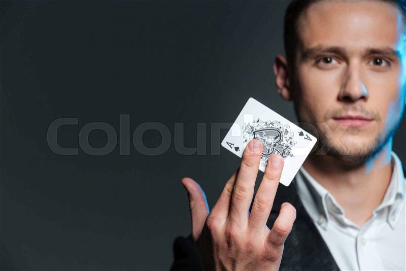 Portrait of handsome young magician holding ace card over grey background, stock photo