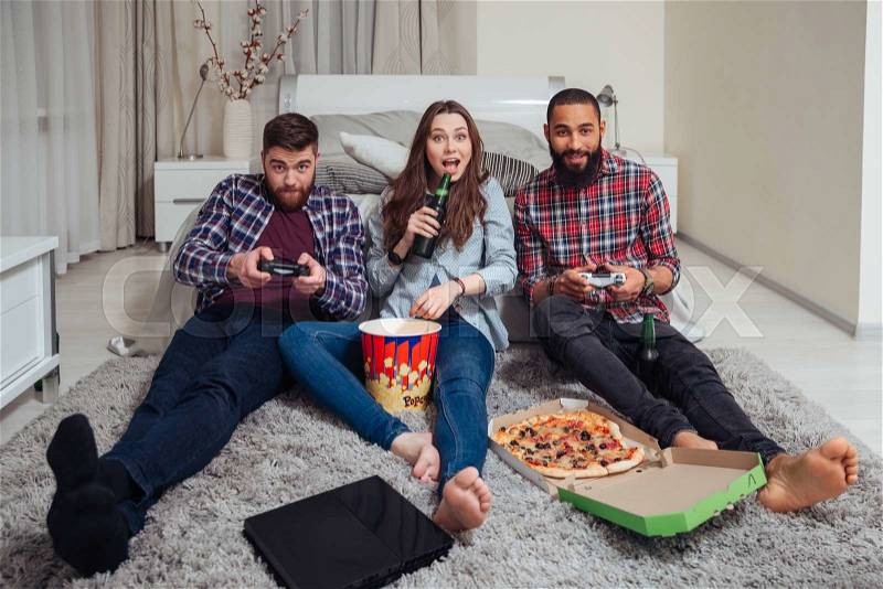 Smiling friends eating pizza and playing computer games at home, stock photo