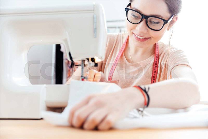 Smiling pretty young woman seamstress sews on the sewing machine in workshop, stock photo