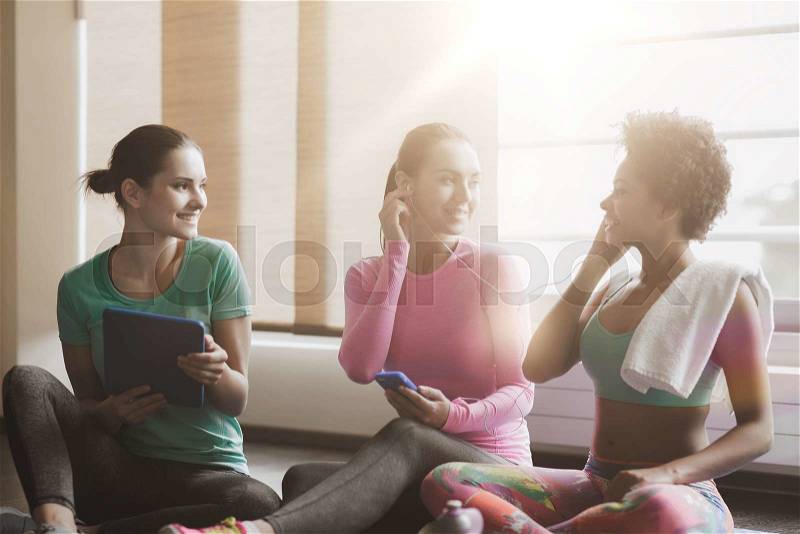 Fitness, sport, technology and lifestyle concept - group of happy women with smartphone, earphones and tablet pc computer listening to music in gym, stock photo