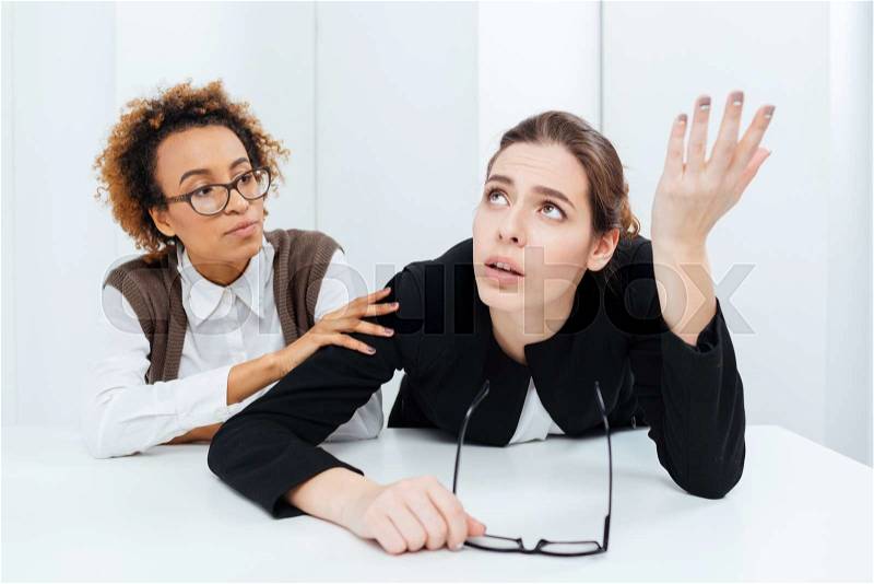 Pretty african american young businesswoman in glasses sitting and supporting her upset despaired colleague in office, stock photo