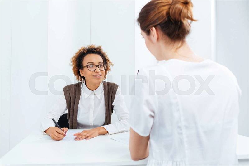 Happy african american young businesswoman in glasses interviewing candidate and writing sitting at the table, stock photo