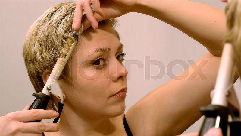 Pretty blond short hair woman curls her hair with curling iron in front of mirror. Beauty and makeup concept, stock photo