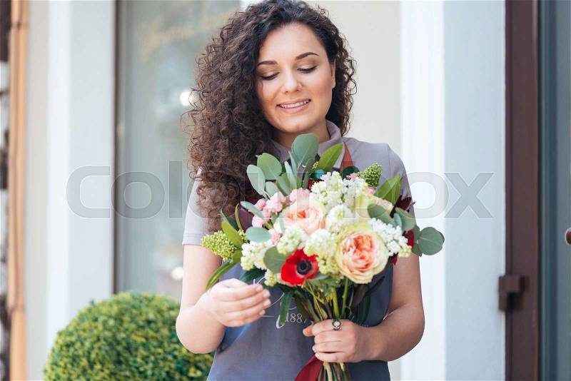 Smiling cute young woman florist with flower bouquet standing in front of shop, stock photo