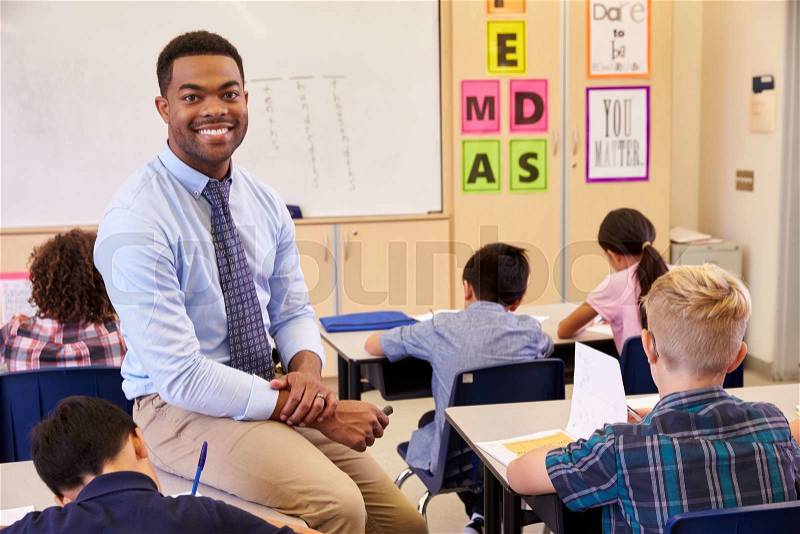 Smiling elementary school teacher sitting on a pupil’s desk, stock photo