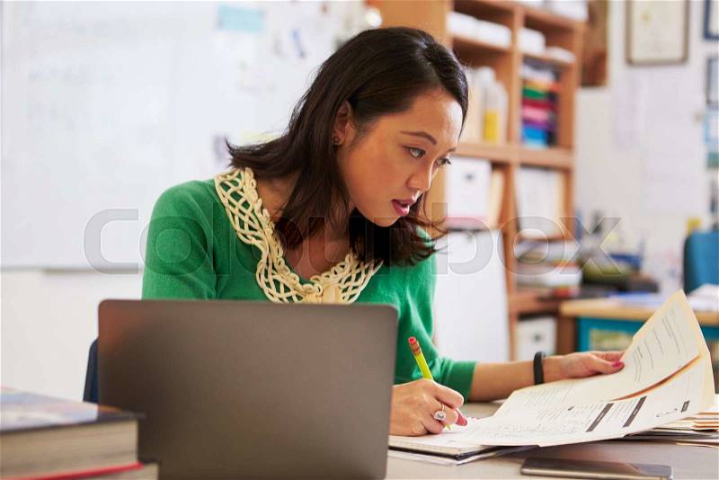 Female Asian teacher at her desk marking students’ work, stock photo
