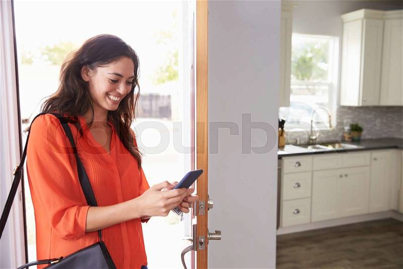 Woman Checking Mobile Phone As She Opens Door Of Apartment, stock photo
