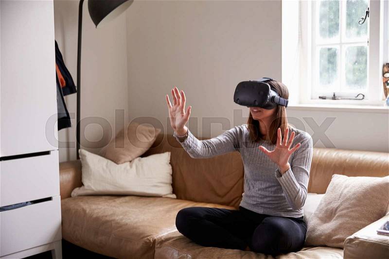 Woman Sits On Sofa At Home Wearing Virtual Reality Headset, stock photo