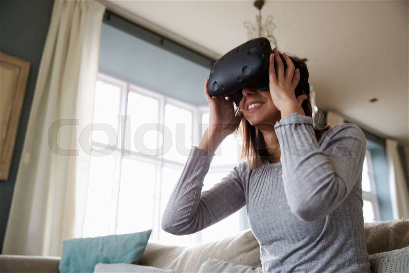 Woman Sits On Sofa At Home Wearing Virtual Reality Headset, stock photo