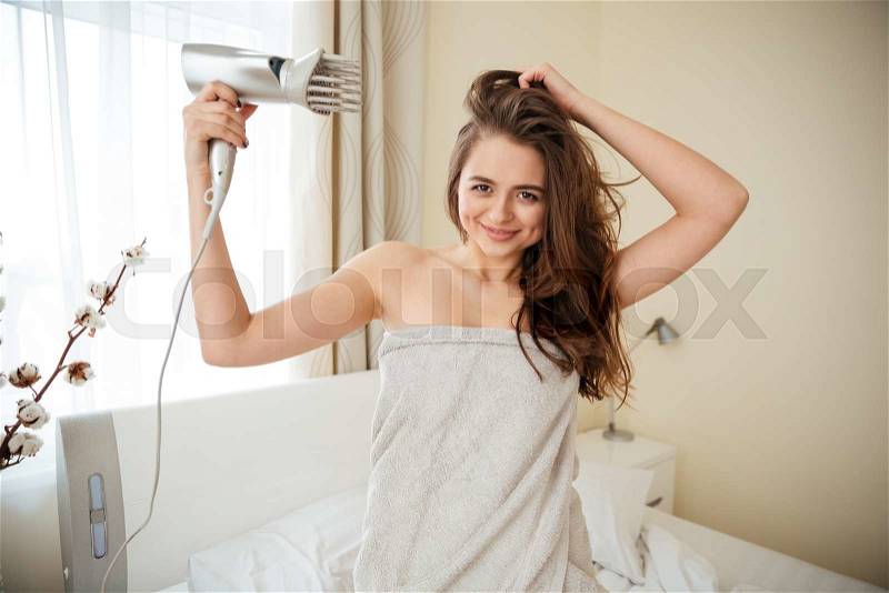 Pretty woman in towel dries hair with hairdryer on the bed at home, stock photo