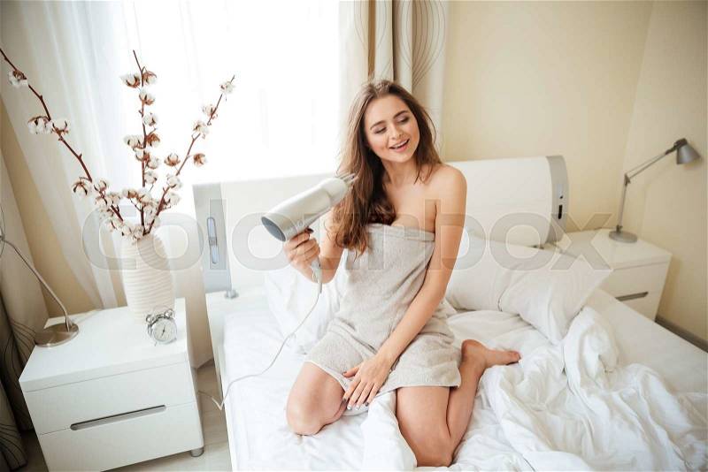 Smiling woman in towel drying hair with hairdryer on the bed at home, stock photo
