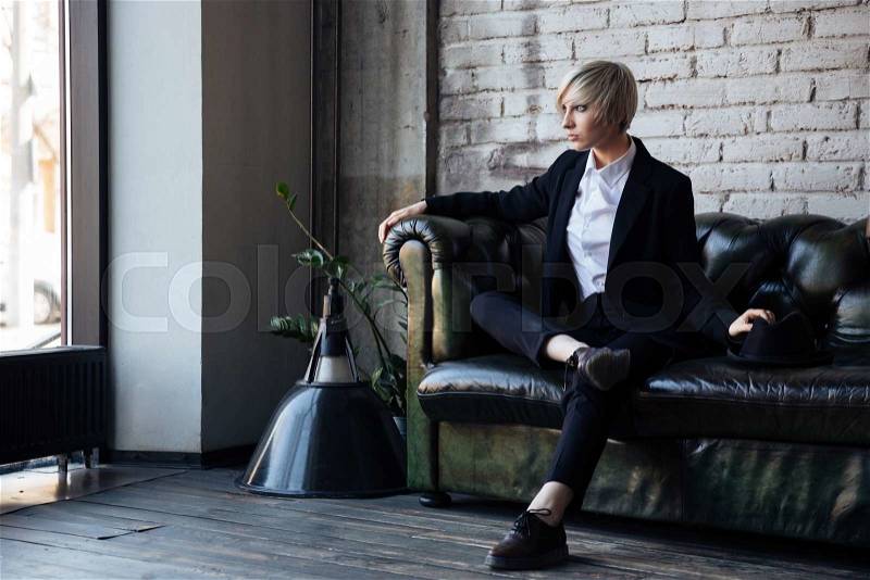 Stylish blonde girl sitting on the leather couch with her legs crossed, stock photo