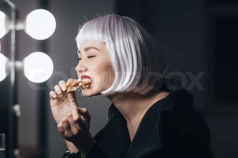 Funny cute young woman in blonde wig eating pizza in dressing room, stock photo