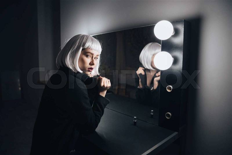 Beautiful young woman in blonde wig and black coat sitting in dressing room, stock photo