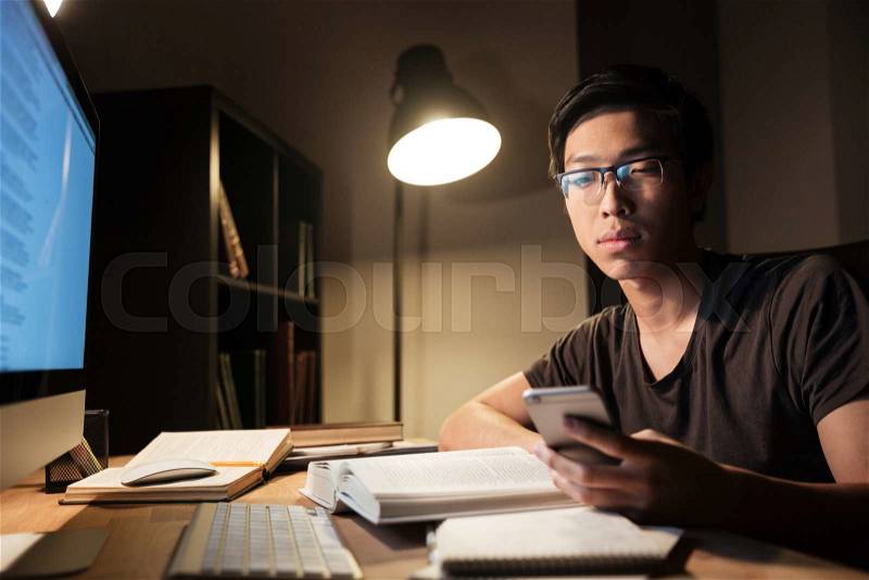 Handsome asian young man in glasses studying and using smartphone in the evening at home, stock photo