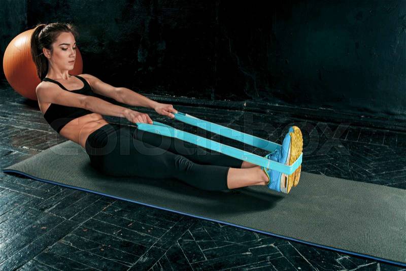 The brunette athletic woman exercising with rubber tape on black wooden floor, stock photo