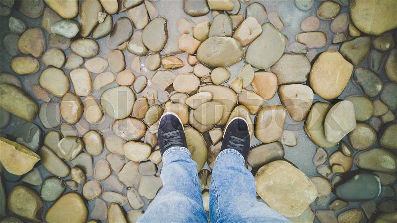 Male feet in sneakers standing on riverbank covered with pebbles, stock photo