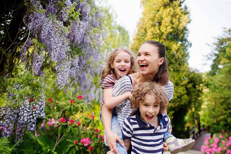 Young woman with her daughter and son on a background of a blossoming garden. They are all very fun. Mom carries a girl on his shoulders. Seven surround flowering plants. Sunny summer day, stock photo