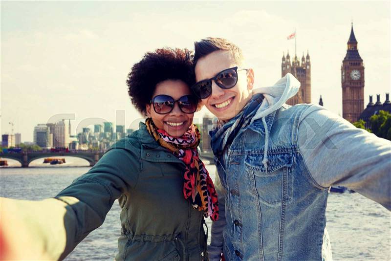 Tourism, travel, people, leisure and technology concept - happy teenage international couple taking selfie over houses of parliament and thames river in london background, stock photo