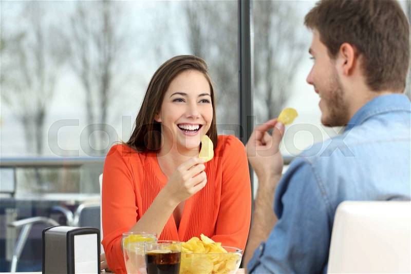 Cheerful couple talking and eating chip potatoes looking each other dating inside a coffee shop with an exterior background, stock photo