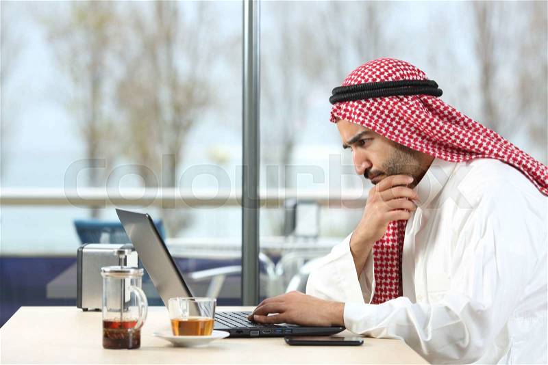 Side view of an arab saudi man worried working with a laptop in a coffee shop interior with the terrace in the background, stock photo