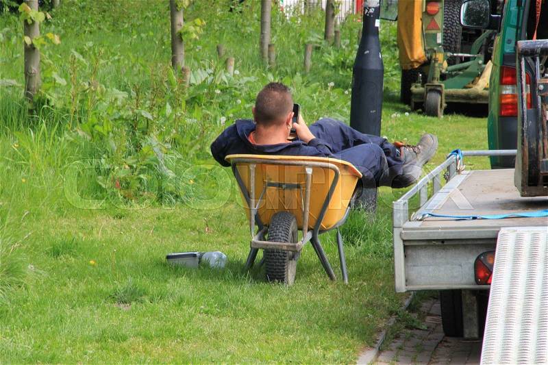 The lazy man in working clothes is playing with his mobile phone in the wheelbarrow in his spare time in the urban park in the city Groningen, stock photo