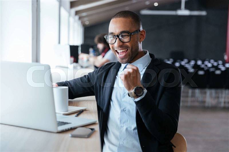 Handsome young businessman sitting at his ddesk, celebrating success with arms raised while looking at his laptop screen, stock photo