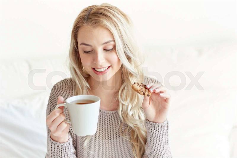 Morning, leisure and people concept - happy young woman with cup of tea eating cookie in bed at home bedroom, stock photo