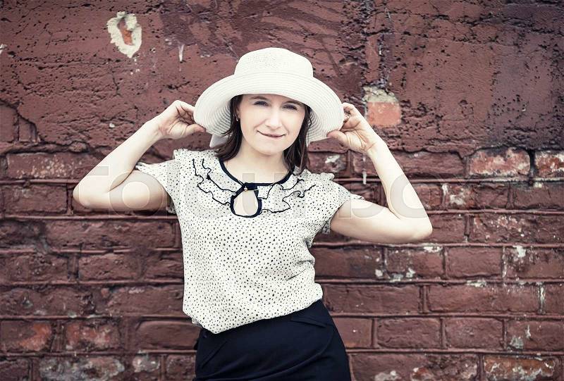 Pretty young woman in white blouse and hat posing against brick wall background. Woman holds the edges of the hat by hands. Toned photo with copy space. Vintage style photo, stock photo