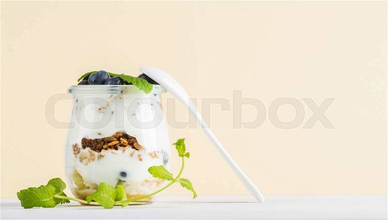 Yogurt oat granola with jam, blueberries and green leaves in glass jar on pastel yellow backdrop, copy space, stock photo